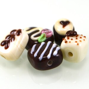 May include: A collection of six glass beads shaped like miniature desserts. The beads are white with brown, pink, and green accents. The beads are designed to look like chocolate-covered strawberries, chocolate squares, and white chocolate with sprinkles.
