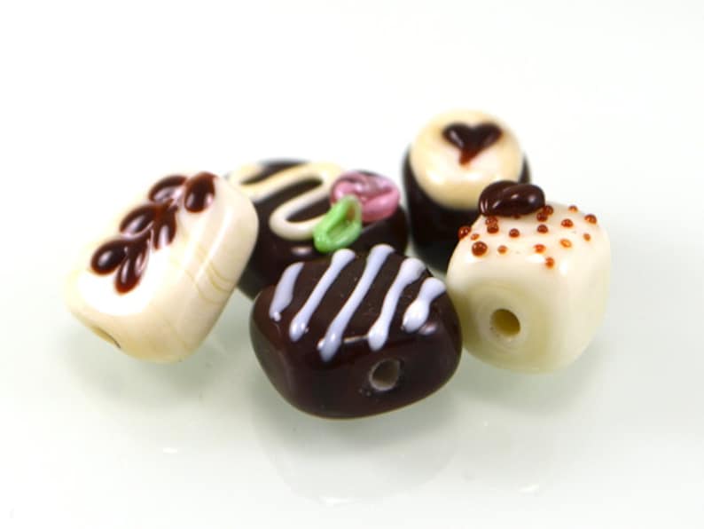 May include: A collection of six glass beads shaped like miniature desserts. The beads are white with brown, pink, and green accents. The beads are designed to look like chocolate-covered strawberries, chocolate squares, and white chocolate with sprinkles.