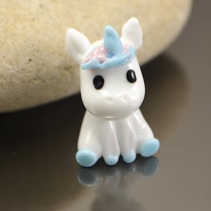 May include: A small white glass unicorn figurine with blue and pink accents. The unicorn has a blue horn, pink ears, and blue hooves.