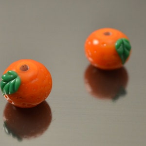 May include: Two small orange glass beads with green leaves. The beads are shaped like oranges and have a small hole in the top.