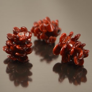 Può includere: Tre perline di pino rosso in vetro. Le perline sono fatte di piccoli pezzi di vetro sovrapposti che ricordano le squame di una pigna.