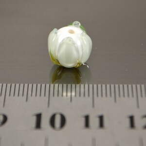 May include: White glass flower bead with green accents. The bead is approximately 1 cm in diameter.