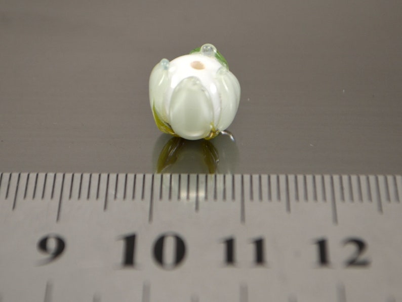 May include: White glass flower bead with green accents. The bead is approximately 1 cm in diameter.