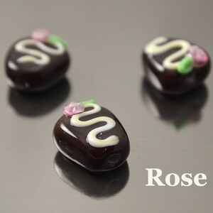 May include: Three small glass ornaments shaped like chocolate candies with white and pink details. The ornaments are on a silver surface. The word "Rose" is printed on the surface.