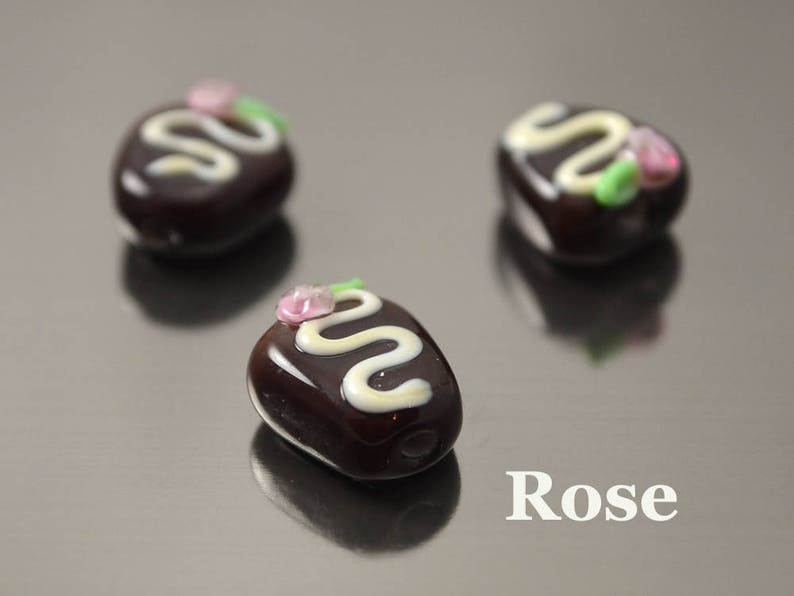 May include: Three small glass ornaments shaped like chocolate candies with white and pink details. The ornaments are on a silver surface. The word "Rose" is printed on the surface.