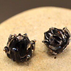 May include: Two handcrafted black glass beads with a unique, textured design. Each bead features a dark, glossy finish with iridescent accents and small, raised details. The beads are displayed on a light-colored, textured surface.