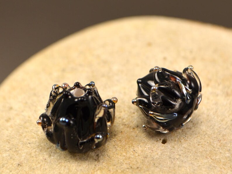 May include: Two handcrafted black glass beads with a unique, textured design. Each bead features a dark, glossy finish with iridescent accents and small, raised details. The beads are displayed on a light-colored, textured surface.