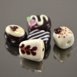 May include: Five glass beads shaped like miniature desserts. The beads are white with brown and white designs. One bead has a swirl design, one has a swirl with a pink flower, one has a brown leaf design, one has brown dots, and one has brown stripes.