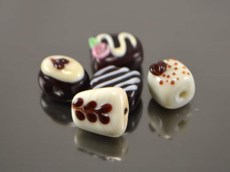 May include: Five glass beads shaped like miniature desserts. The beads are white with brown and white designs. One bead has a swirl design, one has a swirl with a pink flower, one has a brown leaf design, one has brown dots, and one has brown stripes.