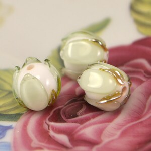 May include: Three white glass beads with a green and gold leaf design. The beads are round and have a hole in the center for stringing.