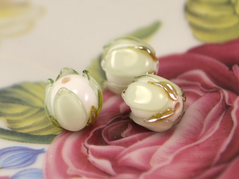 May include: Three white glass beads with a green and gold leaf design. The beads are round and have a hole in the center for stringing.