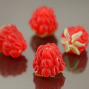 May include: Four red raspberries, each with a slightly different shape and texture, are arranged on a silver surface.