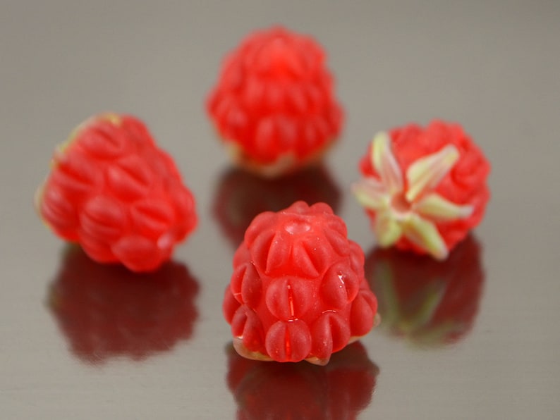 May include: Four red raspberries, each with a slightly different shape and texture, are arranged on a silver surface.