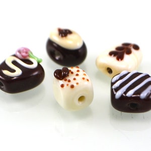 May include: Five glass beads shaped like different chocolate candies. The beads are white and brown with white and brown details.