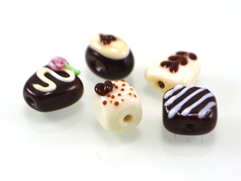 May include: Five glass beads shaped like different chocolate candies. The beads are white and brown with white and brown details.