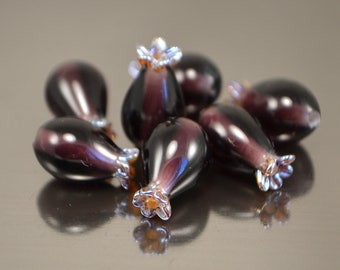 May include: A group of seven small, purple glass ornaments shaped like berries. The ornaments have a shiny, iridescent finish and are arranged in a cluster on a silver surface.