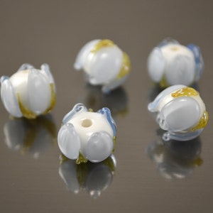 Könnte beinhalten: Fünf weiße Glasperlen mit blauen und goldenen Akzenten. Die Perlen sind blumenförmig mit fünf Blütenblättern.