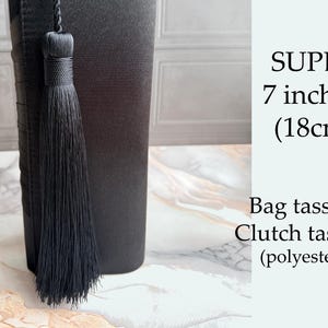 Pode incluir: Borla de saco preta, com 18 cm de comprimento, feita de poliéster. A borla está presa a um objeto preto. O texto à direita diz "SUPER 7 inches (18cm) Bag tassels Clutch tassels (polyester)".
