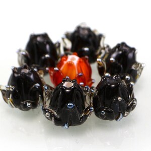 May include: A collection of handmade glass beads in the shape of flowers. The beads are primarily black with silver accents, surrounding a central orange and red bead. The beads are arranged in a circular pattern against a white background.