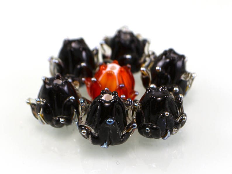 May include: A collection of handmade glass beads in the shape of flowers. The beads are primarily black with silver accents, surrounding a central orange and red bead. The beads are arranged in a circular pattern against a white background.
