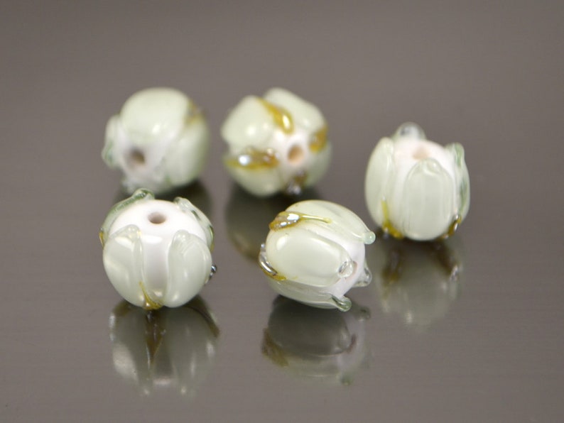 May include: Five white glass beads with a green and gold swirl design. The beads are round and have a slightly irregular shape.