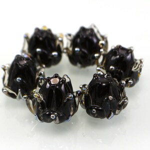May include: A circular arrangement of six black glass frog beads. Each bead has detailed features, including small silver accents and a glossy finish. The frogs are arranged in a circle, creating a decorative element.