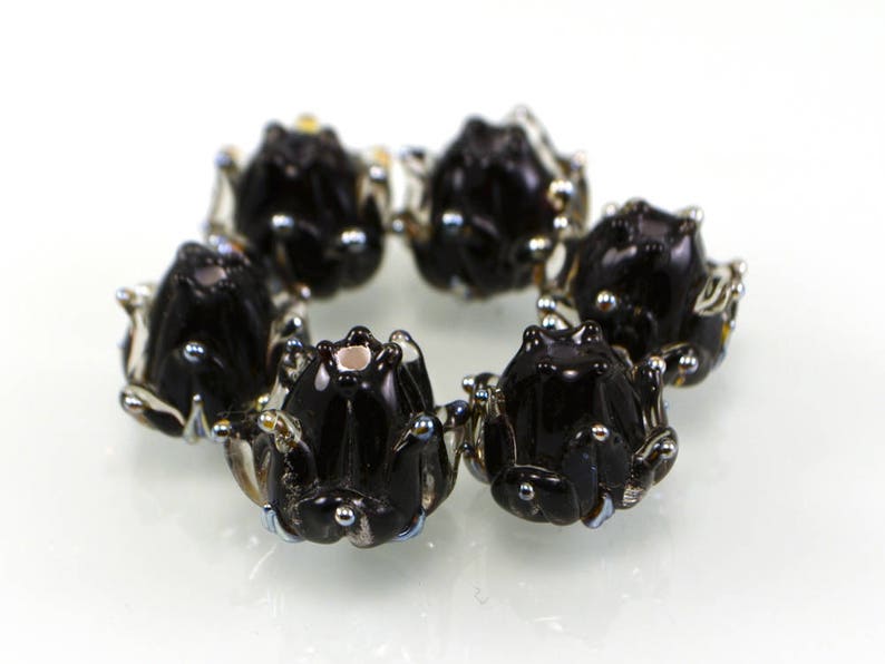 May include: A circular arrangement of six black glass frog beads. Each bead has detailed features, including small silver accents and a glossy finish. The frogs are arranged in a circle, creating a decorative element.