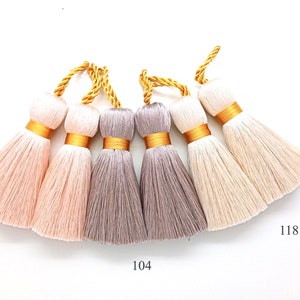 Op de afbeelding: Vijf kwastversieringen in verschillende tinten roze, grijs en wit met gouden accenten. De kwasten zijn geëtiketteerd met de nummers 111, 104 en 118.