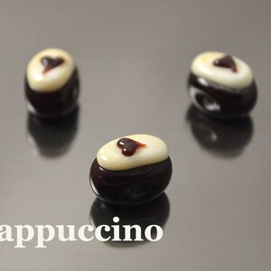 May include: Three dark brown glass beads with a white top and a small brown heart design. The beads are on a reflective surface and the word "Cappuccino" is written in white below the beads.