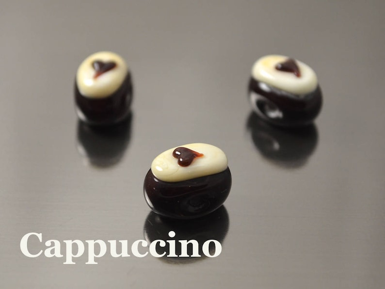May include: Three dark brown glass beads with a white top and a small brown heart design. The beads are on a reflective surface and the word "Cappuccino" is written in white below the beads.