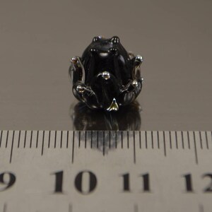 May include: A small, black glass frog figurine with silver accents. The frog is sitting on a reflective surface next to a ruler. The frog's eyes and feet are detailed with silver. The figurine is approximately 1 inch in length.