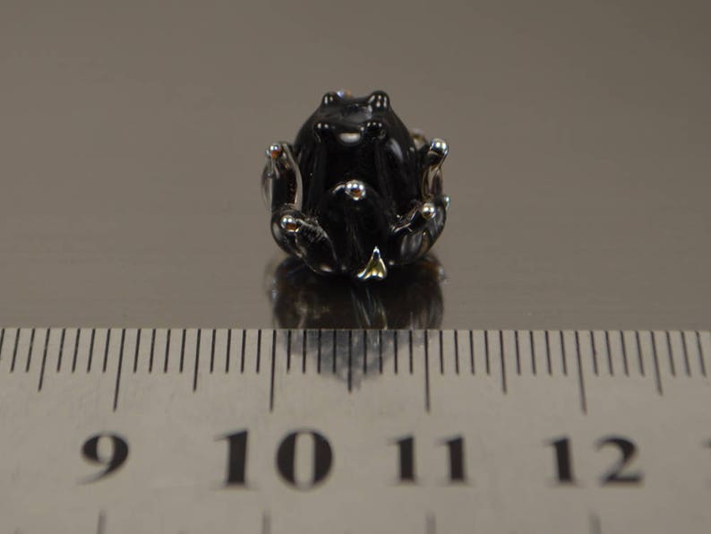 May include: A small, black glass frog figurine with silver accents. The frog is sitting on a reflective surface next to a ruler. The frog's eyes and feet are detailed with silver. The figurine is approximately 1 inch in length.