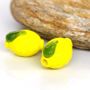 May include: Two yellow glass lemon beads with green glass leaves. The beads are smooth and glossy.