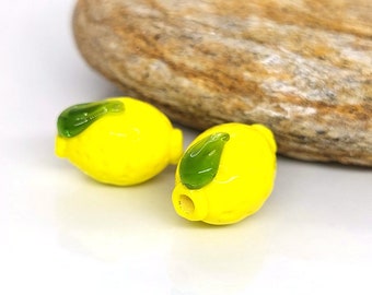 May include: Two yellow glass lemon beads with green glass leaves. The beads are smooth and glossy.