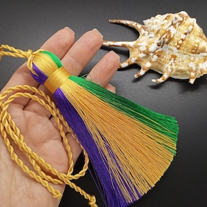 May include: A gold, purple, and green tassel with a gold cord. The tassel is made of soft, fluffy fibers and is tied to a gold cord. The tassel is hanging from a hand.