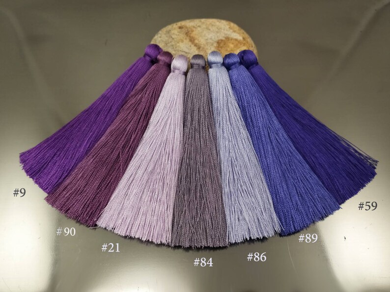 May include: Seven different shades of purple and blue tassels, arranged in a semi-circle on a light brown surface. The tassels are labeled with numbers: #9, #90, #21, #84, #86, #89, and #59.