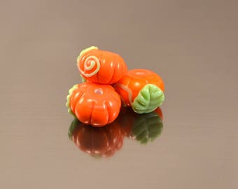 Può includere: Tre perline di vetro arancione a forma di zucca con dettagli verdi e gialli. Ogni perlina ha un motivo a spirale e una foglia verde. Le perline si riflettono su una superficie lucida.