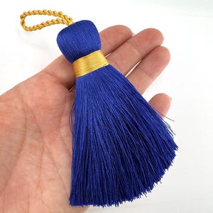 May include: A vibrant royal blue tassel with a gold band and loop. The tassel, made of silky threads, is held in a hand against a white backdrop. This decorative tassel is suitable for various craft projects and measures approximately 15 cm in length.