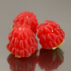 May include: Three red raspberry-shaped candies on a silver surface. The candies have a textured, bumpy surface.