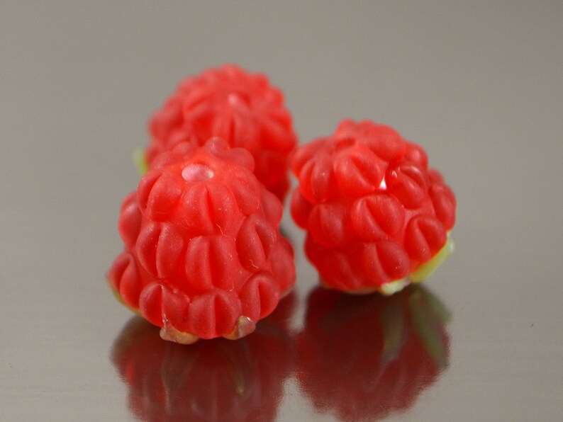 May include: Three red raspberry-shaped candies on a silver surface. The candies have a textured, bumpy surface.