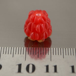 May include: A single, bright red raspberry, approximately 1 cm in diameter, sits on a gray surface next to a ruler.