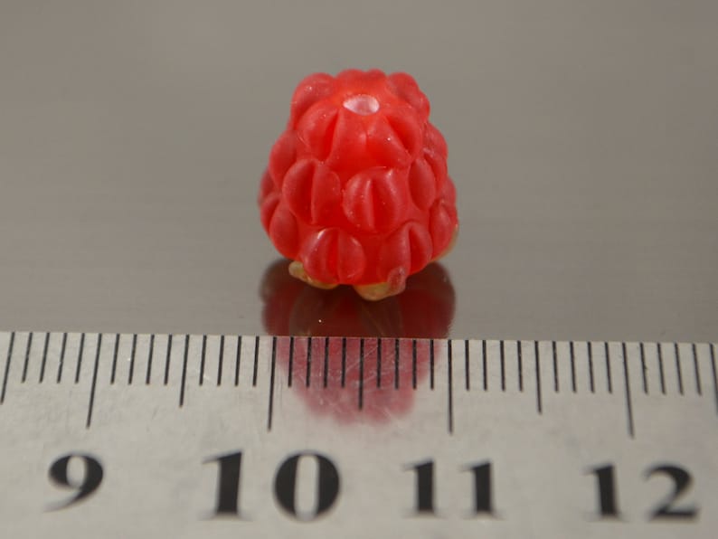 May include: A single, bright red raspberry, approximately 1 cm in diameter, sits on a gray surface next to a ruler.