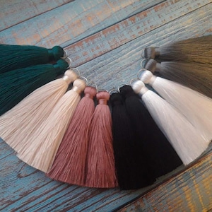 Puede incluir: Un conjunto de diez pares de pendientes de borla en varios colores, incluyendo verde, beige, rosa, negro y blanco. Las borlas están hechas de tela suave y sedosa y están unidas a anillos plateados.