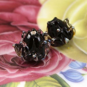 May include: A pair of black glass frog earrings with silver-colored accents. The earrings are detailed with small, round silver beads and sit on a floral patterned background with pink, yellow, and blue hues.