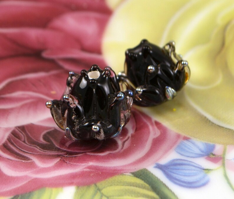 May include: A pair of black glass frog earrings with silver-colored accents. The earrings are detailed with small, round silver beads and sit on a floral patterned background with pink, yellow, and blue hues.