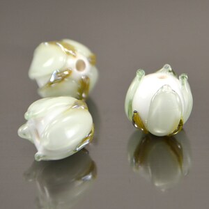 May include: Three white glass beads with green and gold accents, shaped like flowers. The beads are arranged on a reflective surface.