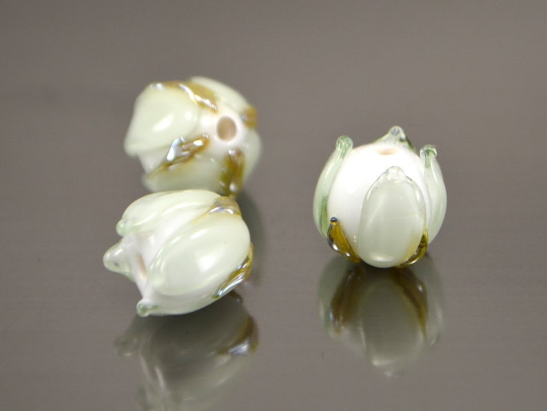 May include: Three white glass beads with green and gold accents, shaped like flowers. The beads are arranged on a reflective surface.