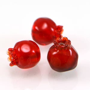 May include: Three red glass pomegranate ornaments. The ornaments are detailed with a textured surface and a crown-like top.