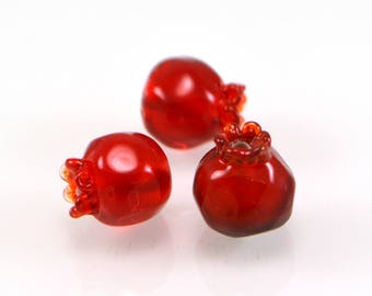 May include: Three red glass pomegranate ornaments. The ornaments are detailed with a textured surface and a crown-like top.