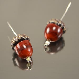 May include: A pair of acorn-shaped earrings with a red and brown glass bead and a silver cap. The earrings are on a white background.